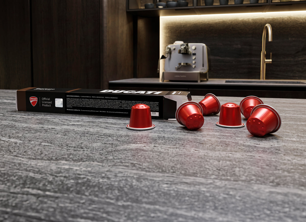 Espresso by Ducati Kapselbox und rote Kapseln auf einer Arbeitsplatte, im Hintergrund die Cuisine Barista M3